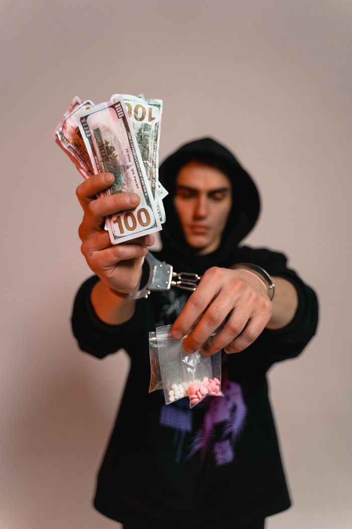 A handcuffed man holds cash and pills, depicting crime and illegal trade.