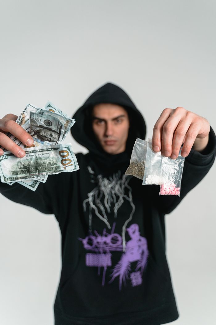About A man in a hoodie holds cash and drug packets, symbolizing crime and illegal trade.
